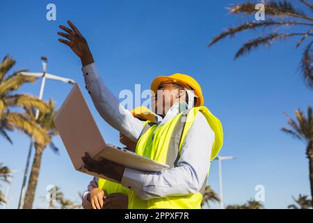 Low angle of young woman builder in uniform and helmet standing and using laptop while discussing project during work Stock Photo