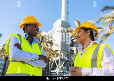 Happy young African American engineers in uniforms and helmets smiling while having conversation against building on sunny day Stock Photo