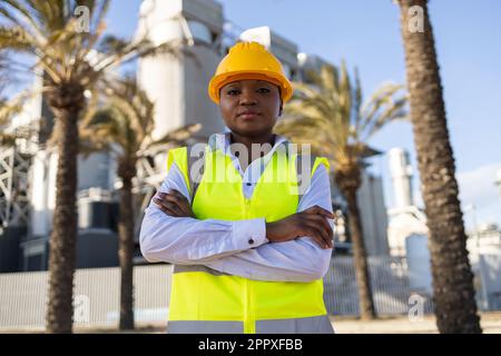 Serious African American female engineer in yellow hardhat and uniform standing with crossed arms and looking at camera while working on construction Stock Photo