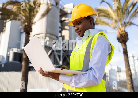 Low angle of young ethnic African America woman builder in uniform and helmet standing and using laptop while looking at project during work Stock Photo