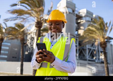 African American female engineer in bright yellow uniform browsing on phone while working construction site and looking at screen with smile Stock Photo