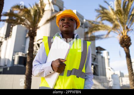 Low angle of young ethnic African America woman builder in uniform and helmet standing holding laptop while looking at project during work Stock Photo