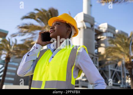 African American female engineer in bright yellow uniform talking on phone while working construction site and looking away with smile Stock Photo