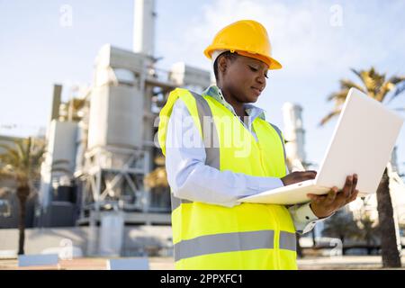 Low angle of young ethnic African America woman builder in uniform and helmet standing and using laptop while looking at project during work Stock Photo