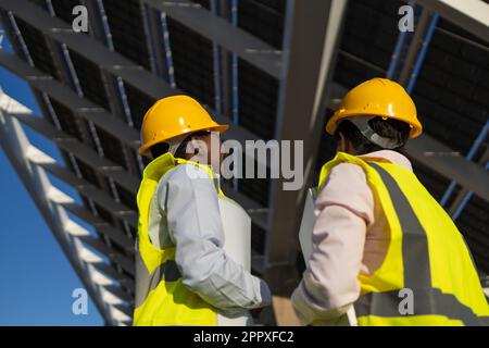 Back view of anonymous engineers in hardhats and uniform standing near installed solar panels on construction site Stock Photo