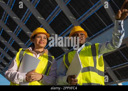 Low angle of young black female engineers in yellow hardhats and vests holding folder with documents and laptop while talking about sustainable solar Stock Photo