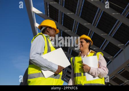 Low angle of young black female engineers in yellow hardhats and vests holding folder with documents and laptop while talking about sustainable solar Stock Photo