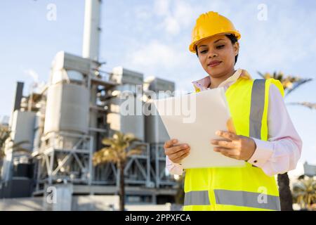 Young ethnic woman in hardhat and engineer vest reading documents while standing on construction site during work process Stock Photo