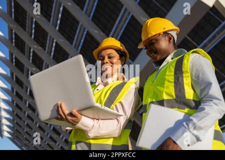 Low angle of young happy African American female builders in hardhat and uniform using laptop while working together and discussing project on constru Stock Photo