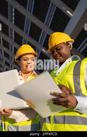 Low angle of young African American women in hardhats and engineer vests smiling and looking at documents while working on construction site Stock Photo