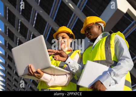 Low angle of young happy African American female builders in hardhat and uniform using laptop while working together and discussing project on constru Stock Photo