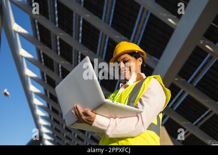 Low angle of happy African American female engineer in yellow vest and helmet standing with laptop while working on construction project with solar ba Stock Photo