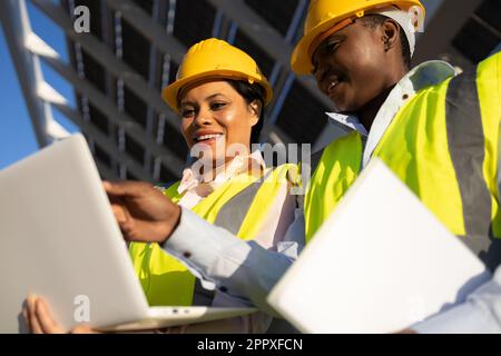 Low angle of young happy African American female builders in hardhat and uniform using laptop while working together and discussing project on constru Stock Photo