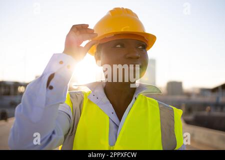 Confident African American woman engineer in hardhat and safety vest touching protective helmet and looking away while standing on street Stock Photo