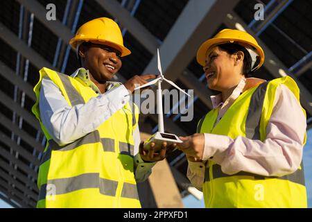 From below cheerful builders in uniforms and helmets using wind turbine while working together on construction site Stock Photo