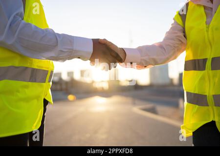 Faceless anonymous engineers in yellow waistcoat uniform shaking hands while standing on city street over blurred landscape with sunlight Stock Photo