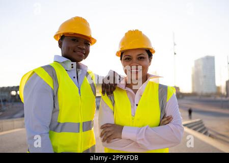 Confident African American female engineers in hardhat and waistcoat looking at camera other while standing on city street Stock Photo