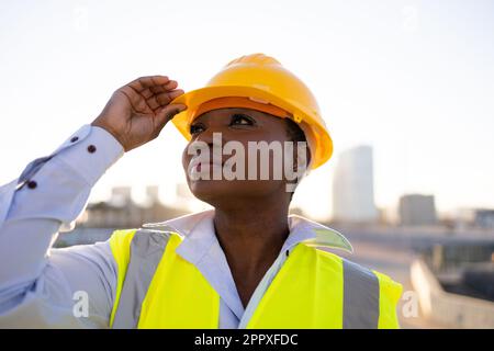 Confident African American woman engineer in hardhat and safety vest touching protective helmet and looking away while standing on street Stock Photo