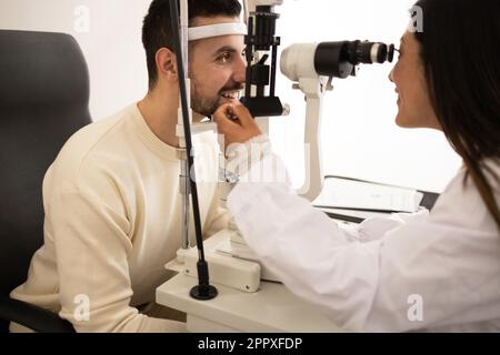 Side view of positive bearded male patient looking at slit lamp while visiting to ophthalmologist for examining vision in medical room Stock Photo