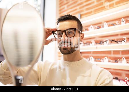 Young male customer choosing eyeglasses while smiling and looking at mirror during visit in optical store Stock Photo