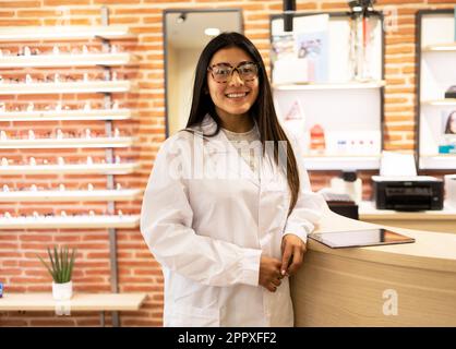 Positive young eye doctor with optical glasses standing near table with tablet and looking at camera while working in optical shop Stock Photo