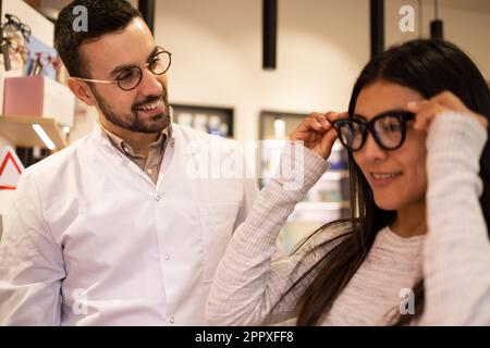 Positive young female applying eyeglasses and standing near male friend while choosing eyewear in optical shop Stock Photo