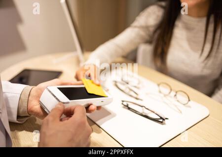 High angle of crop anonymous hand of woman making contactless payment with credit card in optical store while buying new eyewear Stock Photo