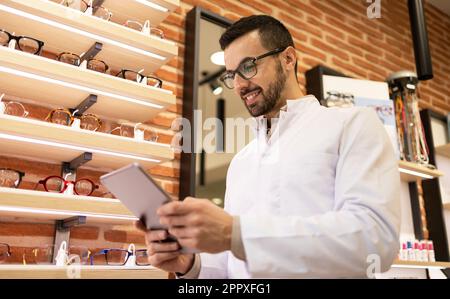 From below portrait of smiling young male optician in white uniform and eyeglasses using digital table while looking at screen in optical shop Stock Photo