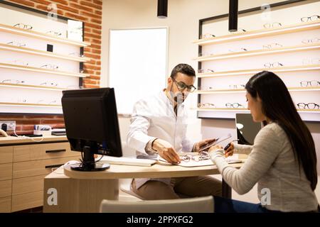 Male optician in white uniform assisting woman in selecting eyeglasses sitting at table near computer monitor at optical shop Stock Photo