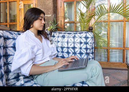 Young concentrated female freelancer sitting on sofa and browsing laptop while resting in cafe during online work in San Miguel de Allende Stock Photo