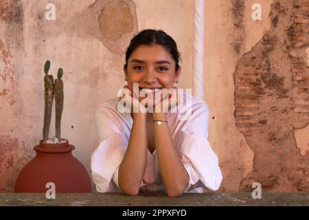 Positive young female in casual clothes smiling and touching chin while sitting at table with green plants and old wall outdoors and looking at camera Stock Photo