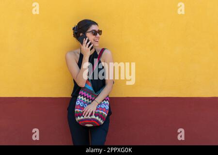 Positive young lady in trendy outfit and sunglasses smiling and looking away while standing against yellow wall and talking on smartphone outdoors in Stock Photo