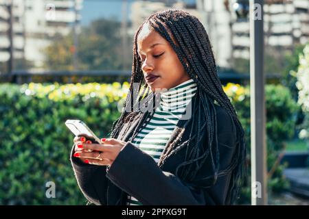 Young African American female in black coat standing on city street and messaging on mobile phone Stock Photo