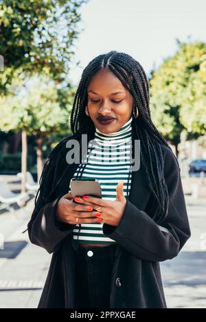 Young African American female in black coat standing on city street and messaging on mobile phone Stock Photo
