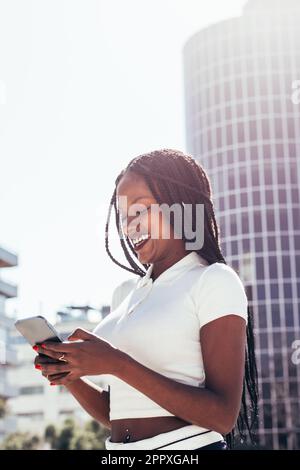Positive African American female in casual clothes and with Afro braids smiling and browsing mobile phone while standing on city street on sunny day Stock Photo