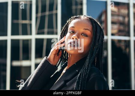Young black female with Afro braids looking at camera while standing near glass wall on city street Stock Photo