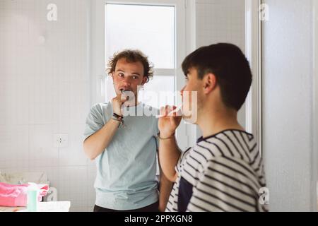 Young gay couple in domestic t shirts brushing teeth in bathroom in morning Stock Photo