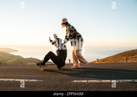 Full body side view of young male with VR goggles sitting on skateboard with outstretched arms while spending time with girlfriend in the seaside coas Stock Photo