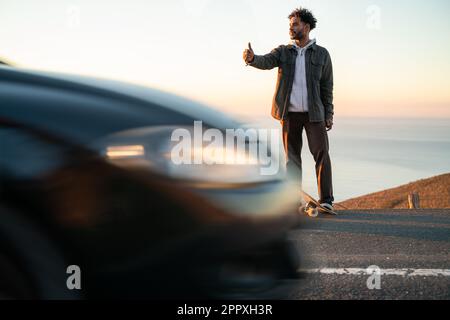 African American male in casual clothes standing with skateboard on road and showing gesture to catch ride while looking away behind driving car durin Stock Photo