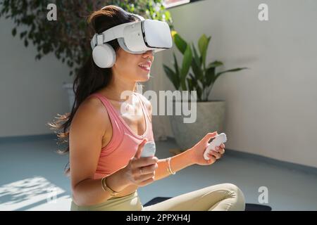 Young asian woman in sportswear sitting on mat and using virtual reality headset while doing fitness exercise during workout Stock Photo