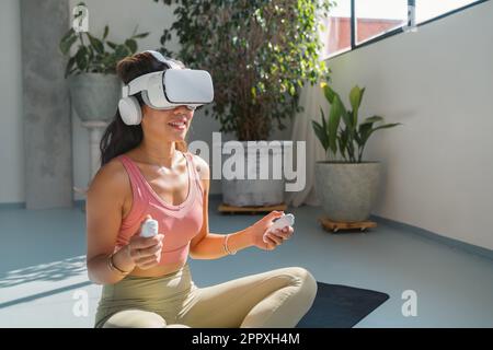 Young asian woman in sportswear sitting on mat and using virtual reality headset while doing fitness exercise during workout Stock Photo