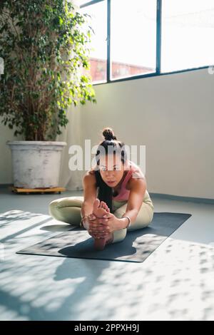 Full body of concentrated young Asian female in sportswear practicing Trianga Mukha Eka Pada Paschimottanasana while stretching on mat during yoga ses Stock Photo