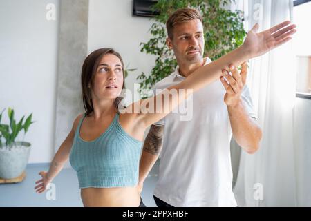 Young male trainer helping female in sportswear doing hands stretching exercise while practicing fitness at studio Stock Photo