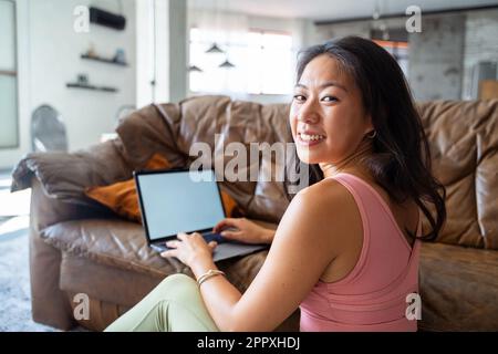 Side view of cheerful ethnic female freelancer in casual clothes smiling and looking at camera over shoulder while sitting on sofa and using laptop in Stock Photo