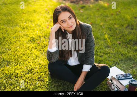 Portrait of attractive cheery businesswoman sitting on grass enjoying good weather holiday pastime outdoors. Sitting woman outdoors. Summer vacation Stock Photo