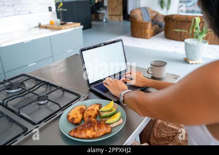 Back view of cropped unrecognizable female freelancer in smart casual clothes working remotely during breakfast in kitchen with cup of coffee using la Stock Photo