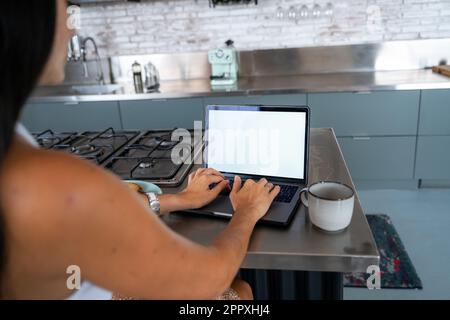Back view of cropped unrecognizable female freelancer in smart casual clothes working remotely in kitchen with cup of coffee using laptop with empty s Stock Photo