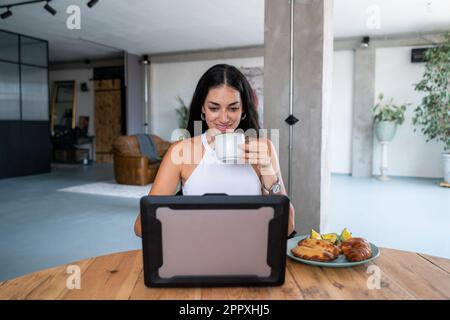 Young cheerful Hispanic female sitting at table drinking hot coffee having empanadas croissant and avocado toasts for breakfast while browsing netbook Stock Photo