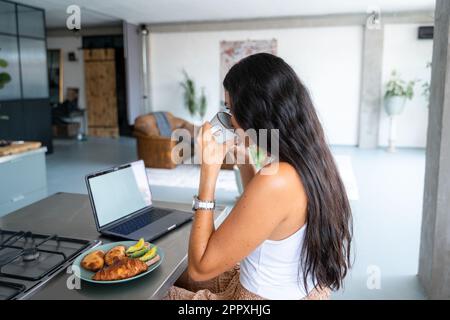 Side view of cropped unrecognizable female freelancer in smart casual clothes working remotely during breakfast in kitchen with cup of coffee using la Stock Photo