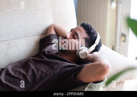 From above relaxed ethnic African American male in casual t shirt lying on comfortable sofa with closed eyes while listening to music on wireless head Stock Photo